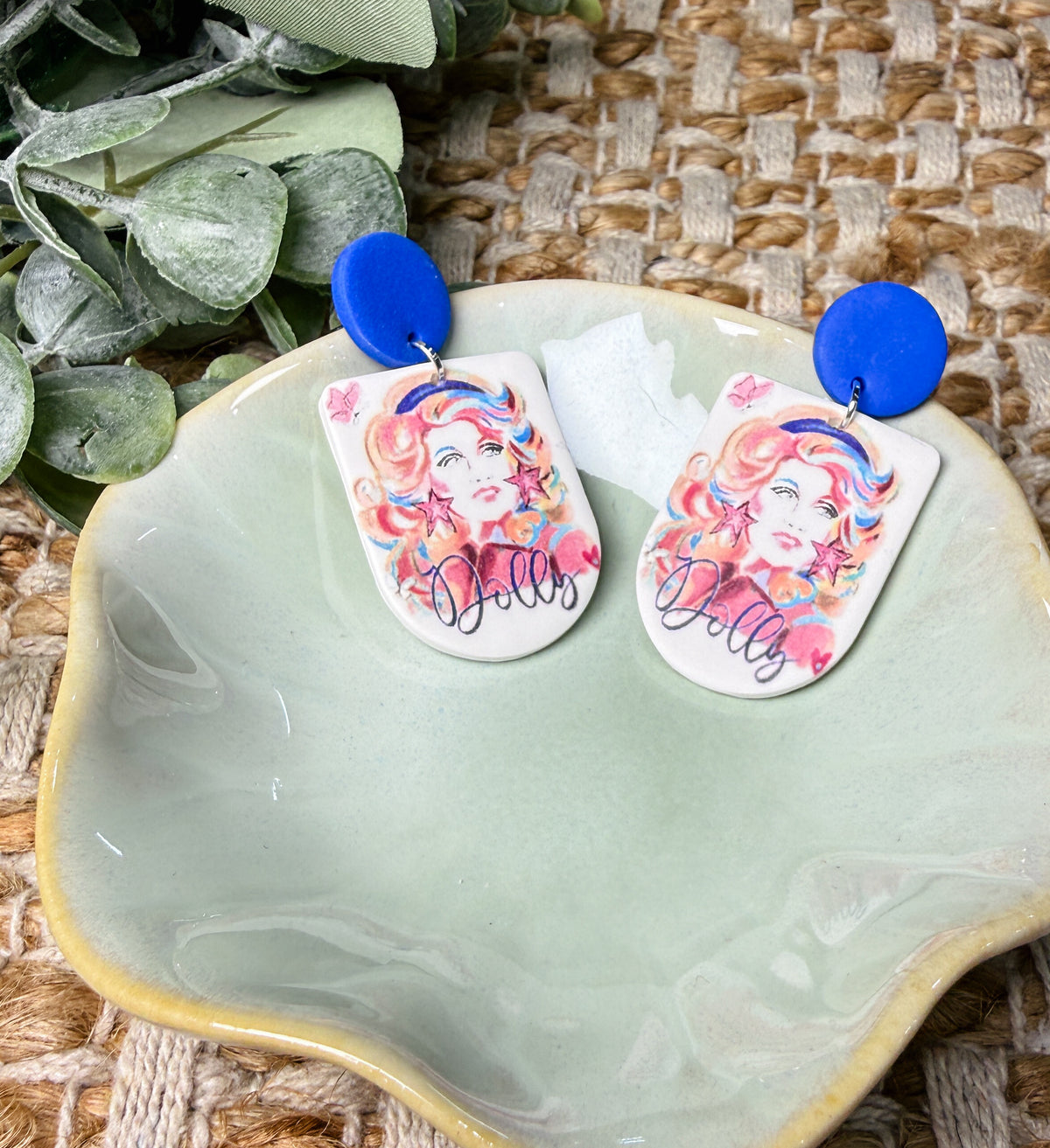 Dolly Parton-inspired dangle earrings featuring colorful retro portrait artwork with pink and orange accents, Dolly lettering, and royal blue round stud tops. Country music statement earrings perfect for concerts and Nashville outfits.