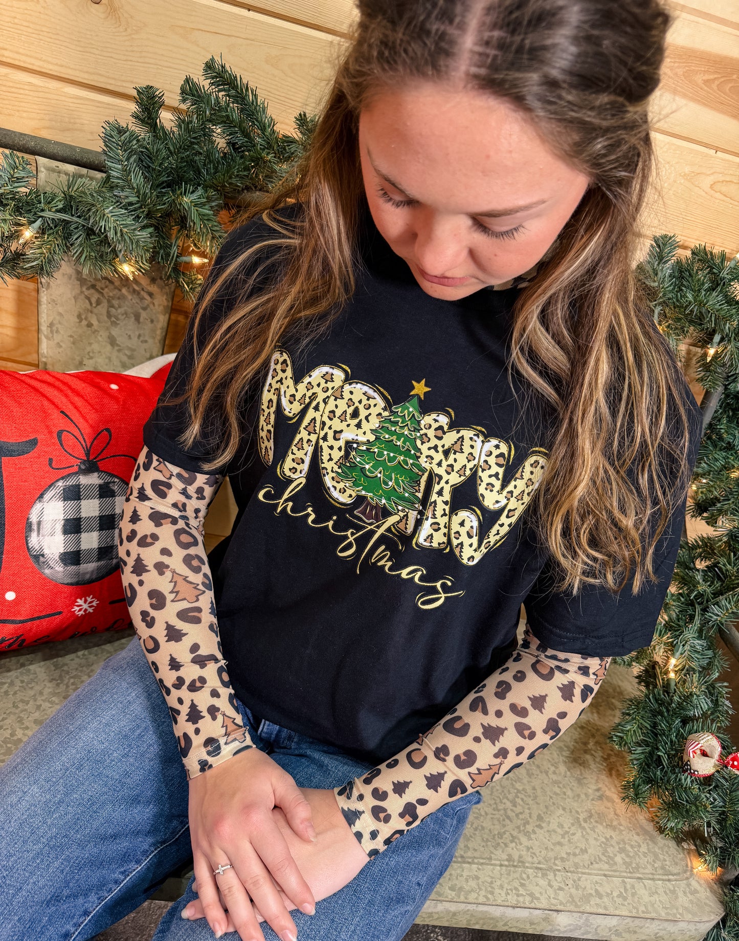 Woman wearing a black Merry Christmas tee layered over a beige mesh long sleeve with leopard and holiday icons, seated in a festive Christmas setting.