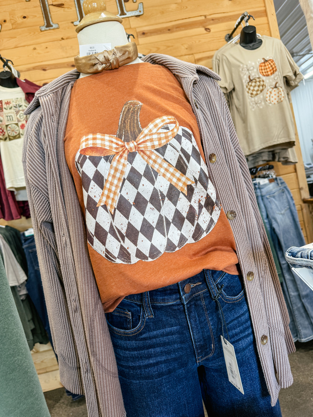 Rust orange fall graphic t-shirt featuring a distressed black and white checkered pumpkin with an orange gingham bow, styled with denim jeans and a corduroy button-up shirt.