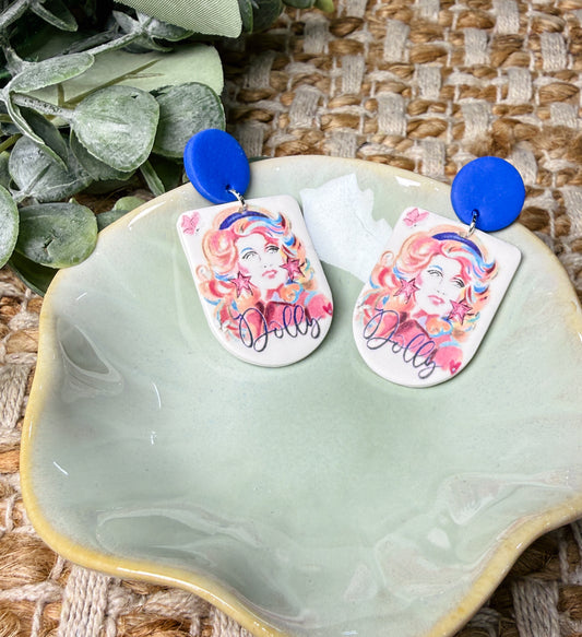 Dolly Parton-inspired dangle earrings featuring colorful retro portrait artwork with pink and orange accents, Dolly lettering, and royal blue round stud tops. Country music statement earrings perfect for concerts and Nashville outfits.