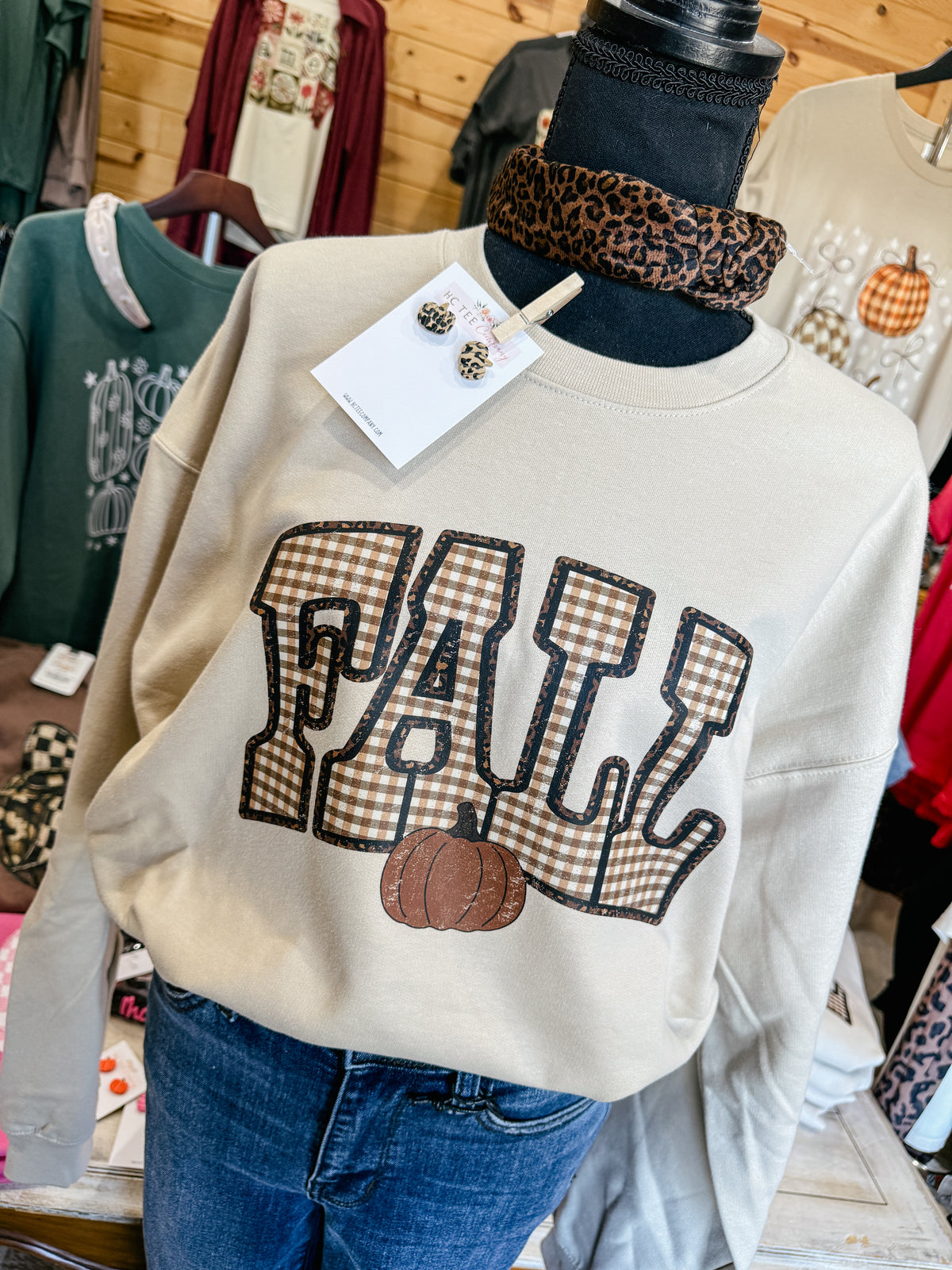 Gildan Sand crewneck sweatshirt featuring distressed brown and cream gingham FALL lettering with a pumpkin graphic, styled with blue jeans and leopard print accessories.