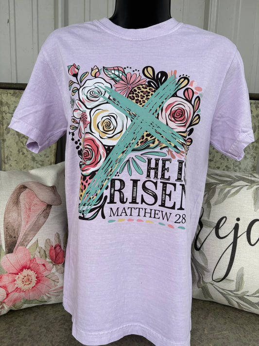 Orchid (light purple) Comfort Colors® t-shirt featuring a colorful floral “He Is Risen” Matthew 28 graphic with a turquoise cross, roses, leopard print accents, and inspirational Christian Easter design. Soft pastel faith-based graphic tee for women.