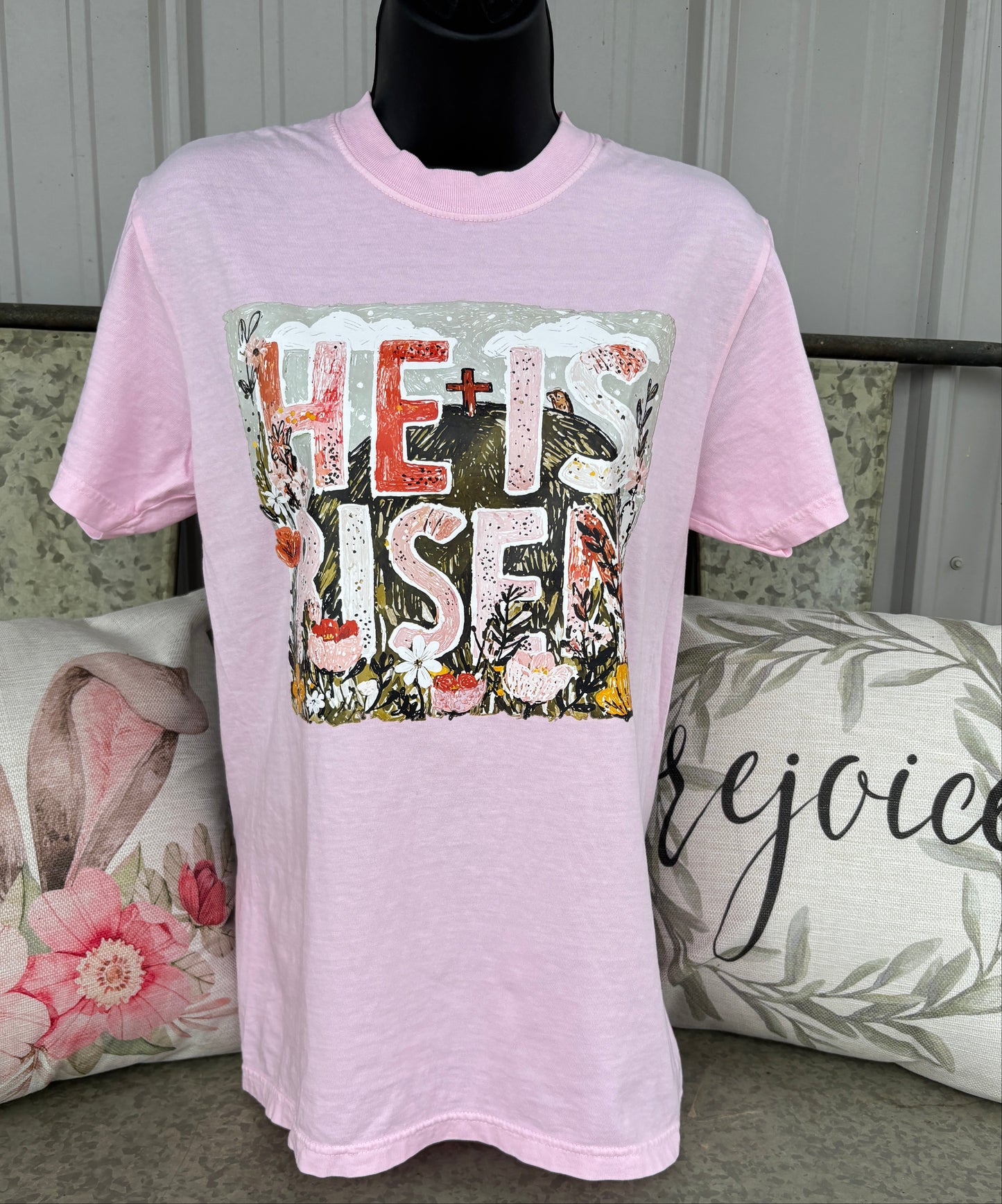 Light pink graphic t-shirt featuring a floral “He Is Risen” Christian design with a cross, flowers, and faith-inspired artwork. Soft pastel Easter tee for women with vintage-style religious graphic and spring boutique aesthetic.