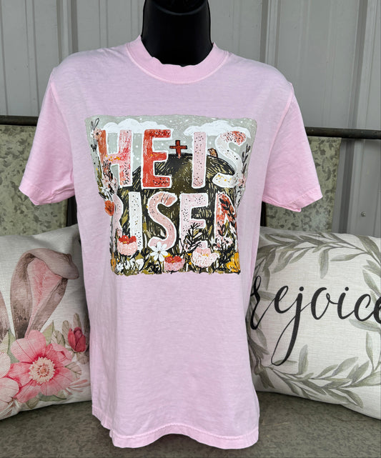 Light pink graphic t-shirt featuring a floral “He Is Risen” Christian design with a cross, flowers, and faith-inspired artwork. Soft pastel Easter tee for women with vintage-style religious graphic and spring boutique aesthetic.