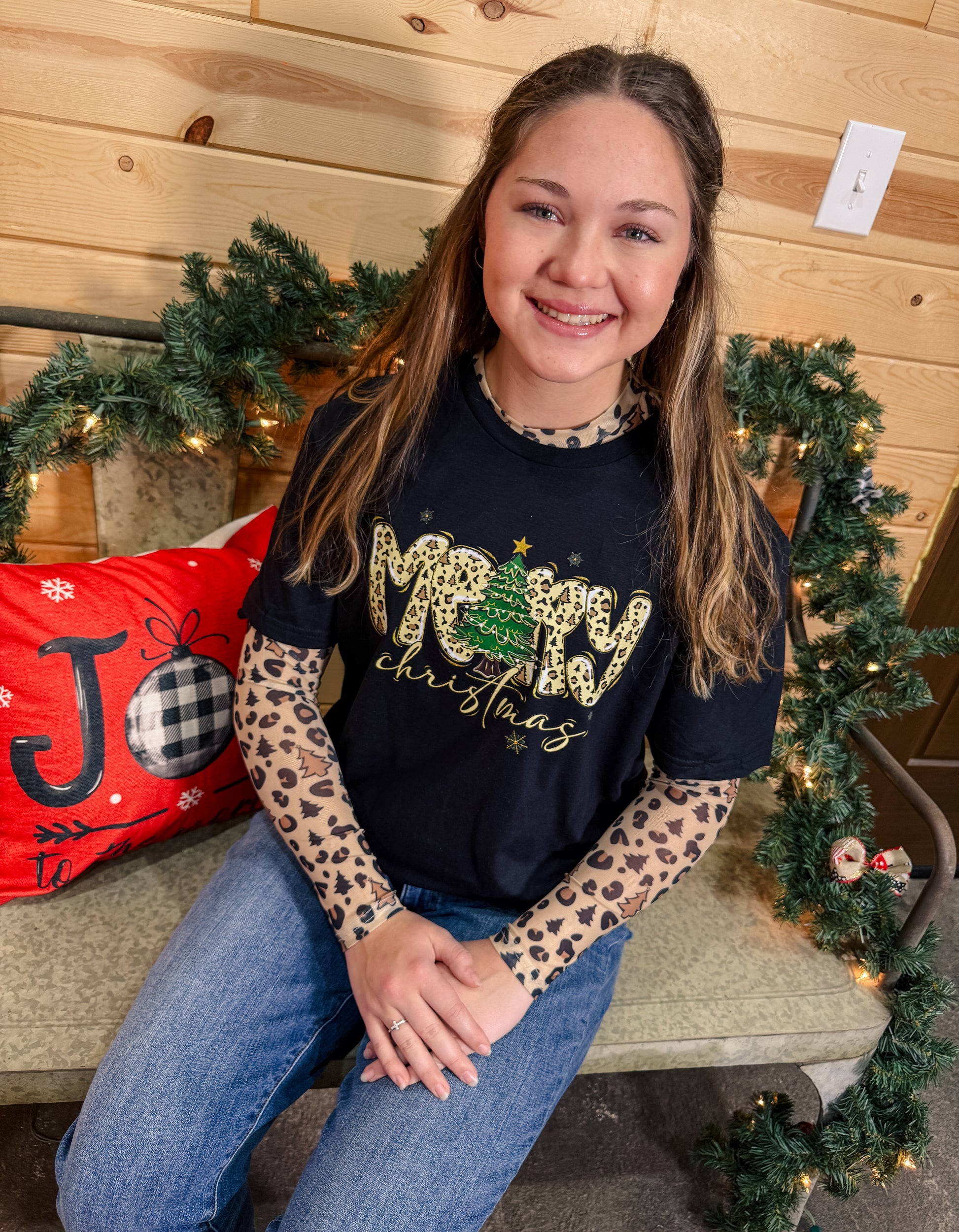 Person wearing a black 'Merry Christmas' shirt with leopard print sleeves, sitting in a festive setting.