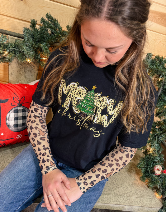 Woman wearing a black Merry Christmas tee layered over a beige mesh long sleeve with leopard and holiday icons, seated in a festive Christmas setting.